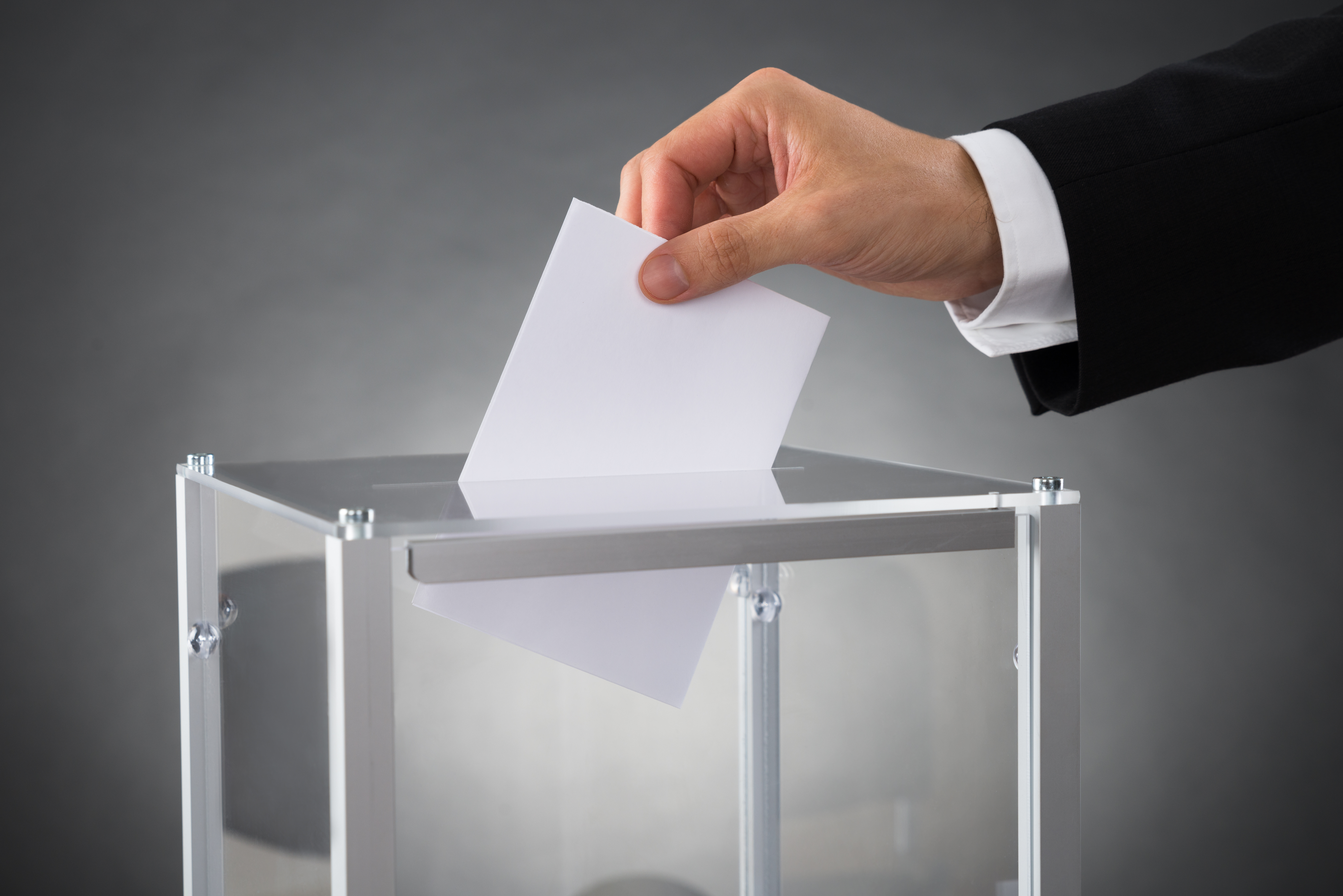 Close-up Of Businessperson Hands Putting Ballot In Box At Desk